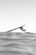 A black and white photograph of a surfer sitting on a surfboard in the ocean, viewed from a low angle with the water surface in the foreground and a pale sky above.