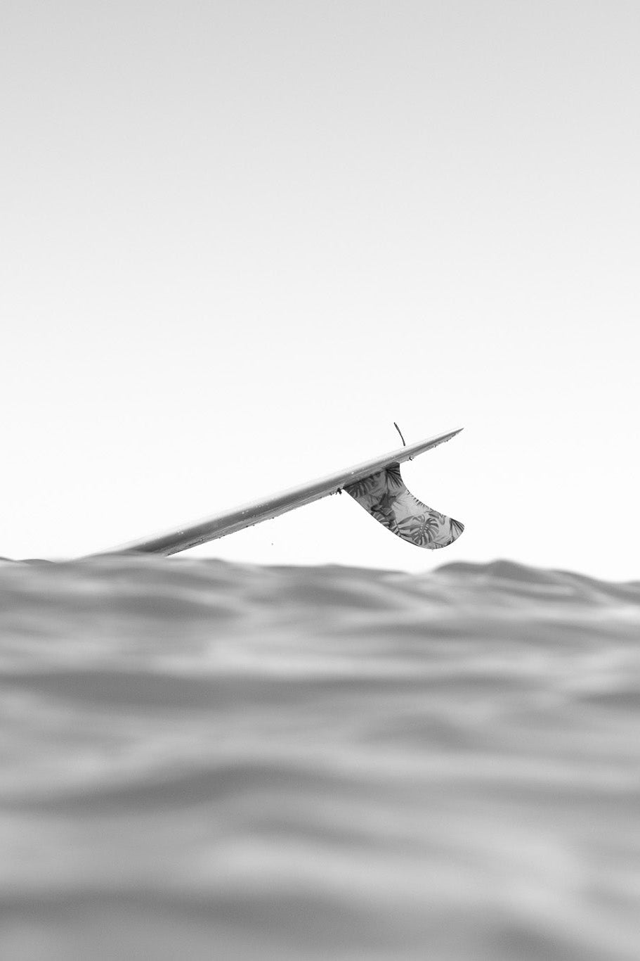 A black and white photograph of a surfer sitting on a surfboard in the ocean, viewed from a low angle with the water surface in the foreground and a pale sky above.